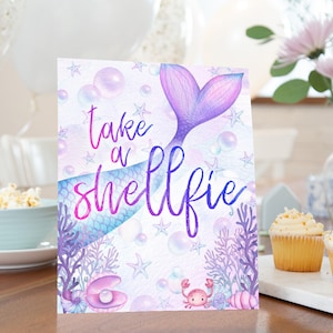 Puede incluir: Un letrero de fiesta con temática de sirenas con la frase "take a shellfie" en una fuente caprichosa. El letrero presenta una cola de sirena morada y azul, burbujas, estrellas de mar y coral. La paleta de colores incluye tonos de morado, azul y rosa.