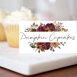 Op de afbeelding: Een witte kaart met de tekst "Pumpkin Cupcakes" in elegant schrift, omlijst door aquarel bloemstukken in bordeauxrood, bruin en goud. Cupcakes met witte glazuur staan op de achtergrond.