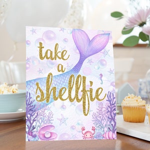 Puede incluir: Un letrero con la frase "take a shellfie" en letras doradas, con una cola de sirena, burbujas, estrellas de mar y coral en tonos morados, azules y rosas. El letrero está sobre una mesa con cupcakes y decoraciones.