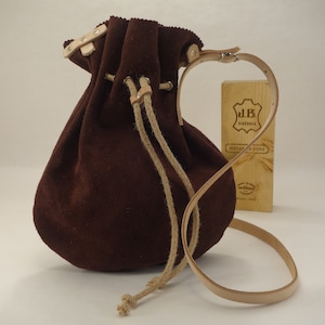 May include: A brown suede drawstring bag with a tan leather strap and a tan rope drawstring. The bag is sitting on a white surface next to a wooden box with the text "J.B. RIOJENZA JHOTAS Y VINO" printed on it.
