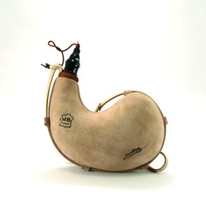 Op de afbeelding: Een bruine leren fles met een bruine leren riem en een zwarte stop. De fles heeft een label met de tekst "J.B. Barcelona" en een handtekening aan de zijkant.