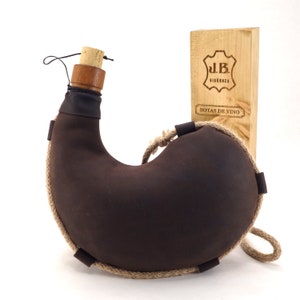 May include: A brown leather wine bottle with a cork stopper and a rope handle. The bottle is sitting on a white surface next to a wooden box with the text "J.B. BIGUENZA BOTAS DE VINO" printed on it.