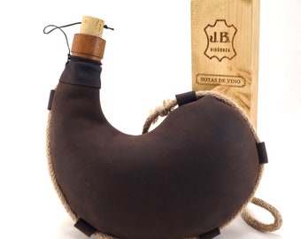 May include: A brown leather wine bottle with a cork stopper and a rope handle. The bottle is sitting on a white surface next to a wooden box with the text "J.B. BIGUENZA BOTAS DE VINO" printed on it.