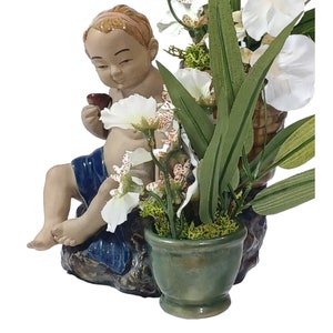 May include: A ceramic figurine of a child sitting with a cup in hand, surrounded by white and pink flowers in a green ceramic pot. The figurine is wearing a blue and white patterned cloth.