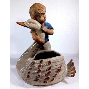 May include: A ceramic planter shaped like a swan with a boy figurine holding onto the swan's neck. The swan has brown and white feathers and a brown beak. The boy is wearing a blue shirt and white pants.