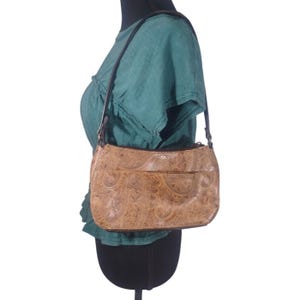 May include: Tan shoulder bag with a paisley pattern. The bag has a front pocket and a black strap. The bag is worn over a green top.