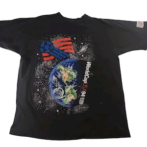 May include: Black t-shirt featuring a graphic of Earth, a soccer ball, and a stylized American flag. The text "WorldCup USA 94" is printed on the right side of the globe. The shirt has a crew neck and short sleeves.