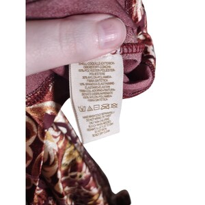 May include: A close-up of a tag with text in multiple languages, including English, French, and Spanish. The tag is attached to a maroon fabric with a floral pattern in shades of gold and brown. The tag provides fabric composition and care instructions.