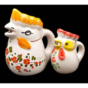 May include: Two white ceramic pitchers shaped like chickens, featuring yellow and orange crests, orange beaks, and hand-painted red floral patterns with green leaves. The larger pitcher has a handle on the right, while the smaller one is also handled.