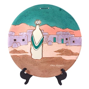 May include: A ceramic plate with a painted scene of a woman in a white dress with green accents standing in front of a village with pink and purple buildings. The plate is on a black stand and signed "Vera Russell".