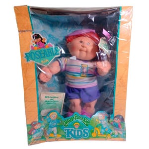 May include: A Cabbage Patch Kids poseable soft doll with red hair and a purple hat, wearing a striped shirt and purple shorts. The doll is inside a cardboard box with orange borders, featuring the text Poseable Soft and Birth Certificate.
