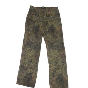 May include: Camouflage-patterned pants in shades of green, brown, and tan. The pants have a straight leg cut, a button and zipper closure, and pockets. A small orange logo is visible near the right pocket.