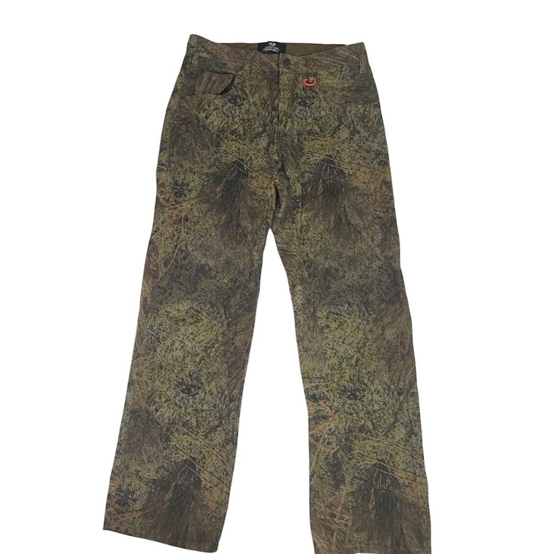 May include: Camouflage-patterned pants in shades of green, brown, and tan. The pants have a straight leg cut, a button and zipper closure, and pockets. A small orange logo is visible near the right pocket.