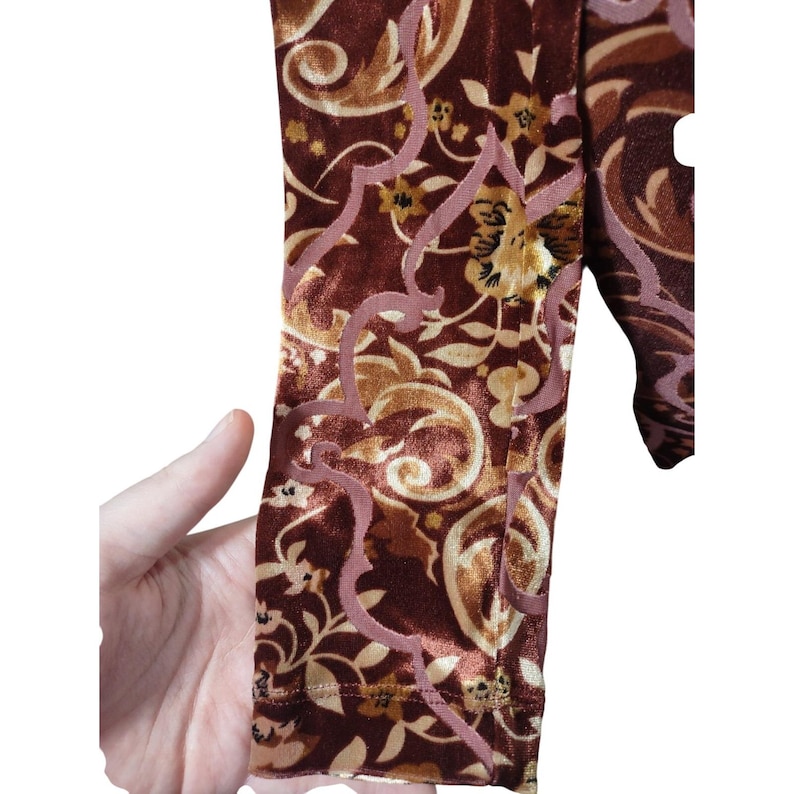 May include: Close-up of a section of fabric with a rich, velvety texture. The fabric features a complex pattern of swirling floral and scroll designs in shades of brown, gold, and mauve. The design is detailed and intricate.