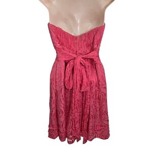May include: A strapless, knee-length pink dress. The dress features a textured bodice and a bow at the waist. The skirt has a subtle floral pattern. The dress is displayed on a mannequin.