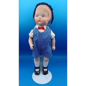 May include: A vintage doll stands on a white stand, wearing a blue polka-dot romper, white shirt, and a red bow tie. The doll has a matching blue bonnet and black shoes. The doll is a collectible item.