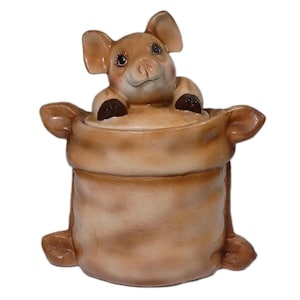 May include: A ceramic pig-shaped cookie jar. The pig is light brown with dark brown hooves and is peeking out of a tan sack. The pig has detailed facial features, including eyelashes and a smiling expression. The jar is designed to look like a sack.