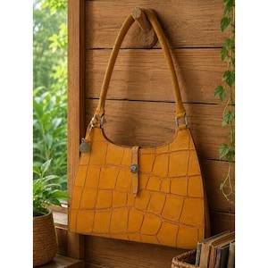 May include: A mustard yellow shoulder bag with a textured crocodile skin pattern. The bag features a single strap, a central vertical leather tab with a silver-toned metal turn-lock closure, and a small circular silver-toned metal charm attached to the side.