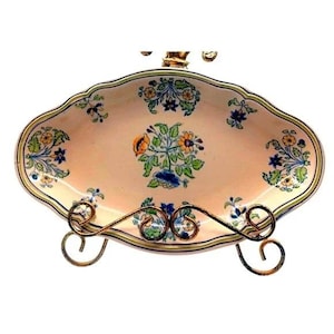 May include: An oval-shaped, antique-style serving dish with a floral design in shades of blue, yellow, and orange on a pale pink background. The dish is supported by a decorative, ornate metal stand. The rim is trimmed with a green and yellow border.