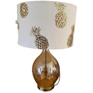 May include: A decorative table lamp with a gold-toned glass base and a white lampshade. The lampshade features a repeating pattern of gold sequined pineapples. The lamp has a brass-colored base and a curved handle.