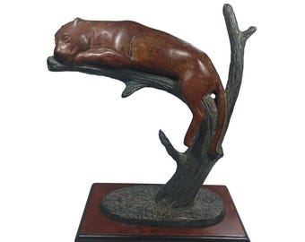 May include: A bronze sculpture of a brown cougar resting on a tree branch. The sculpture is mounted on a wooden base.