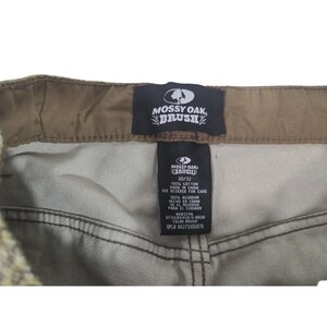 May include: Close-up of the waistband and label of Mossy Oak Brush pants. The label indicates a 30/32 size and 100% cotton material. The pants are a neutral color, with a brown waistband and stitching.