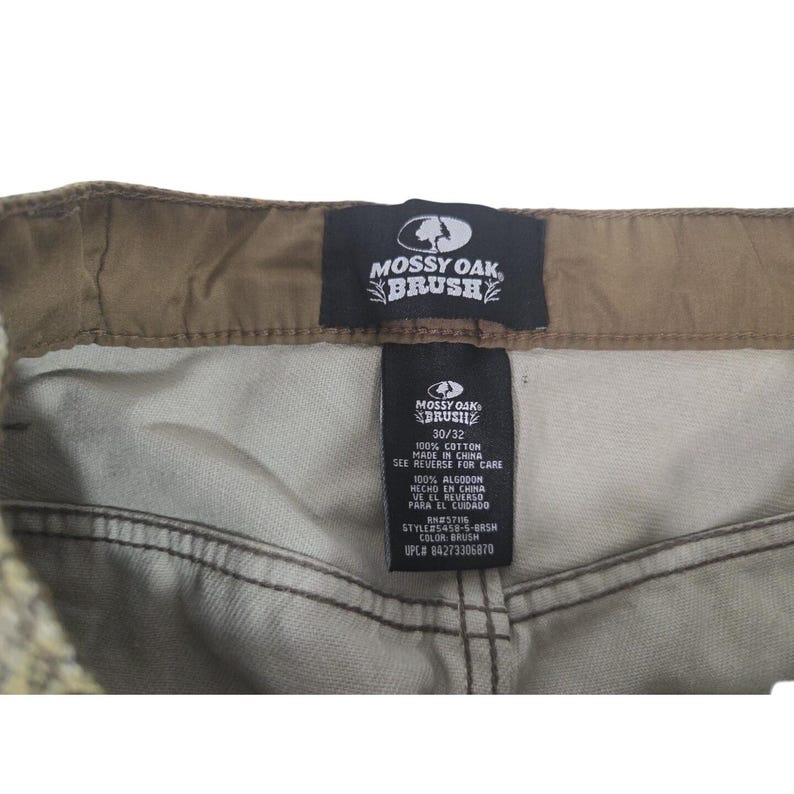 May include: Close-up of the waistband and label of Mossy Oak Brush pants. The label indicates a 30/32 size and 100% cotton material. The pants are a neutral color, with a brown waistband and stitching.