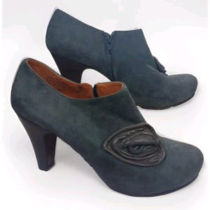 May include: A pair of dark blue suede heeled ankle boots with a decorative eye design on the front of one boot. The boots have a zipper on the side and a chunky heel.