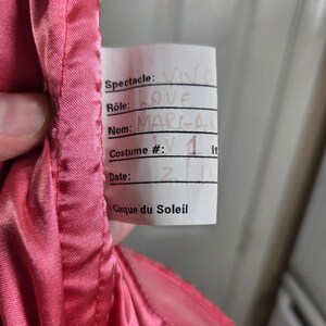 May include: A close-up of a pink costume with a tag. The tag reads "Spectacle: VIVA LOVE, Nom: MARGA, Costume #: 1, Cirque du Soleil". The costume is made of a shiny, silky material.