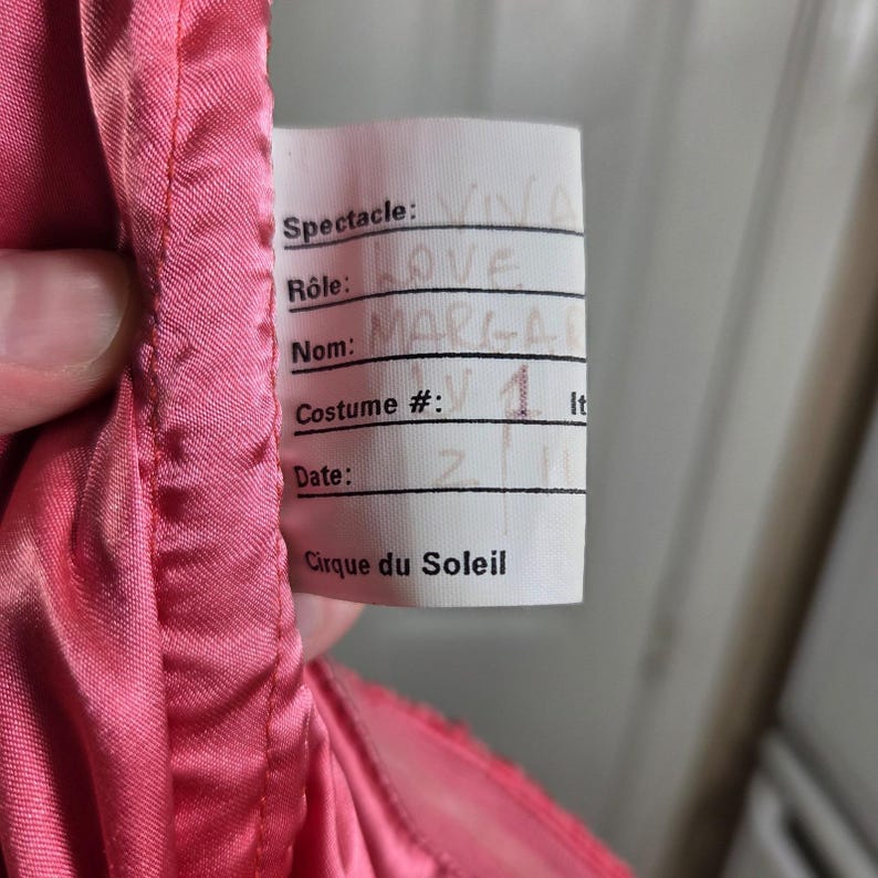 May include: A close-up of a pink costume with a tag. The tag reads "Spectacle: VIVA LOVE, Nom: MARGA, Costume #: 1, Cirque du Soleil". The costume is made of a shiny, silky material.