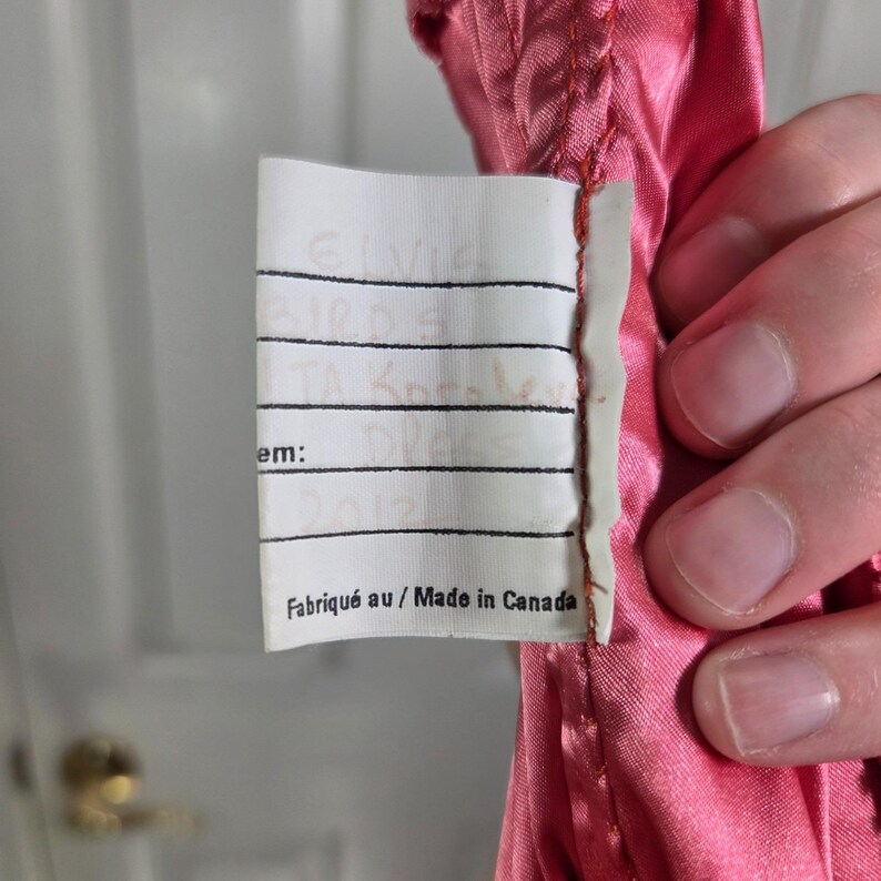 May include: A close-up of a pink satin fabric with a white tag attached. The tag has handwritten text and the words "Fabriqu&eacute; au / Made in Canada" printed at the bottom.