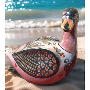 May include: A decorative ceramic duck figurine with a red, black, and white color scheme. The duck has intricate patterns and designs on its wings and body, with a detailed beak and head. The figurine is set on a sandy beach with the ocean in the background.