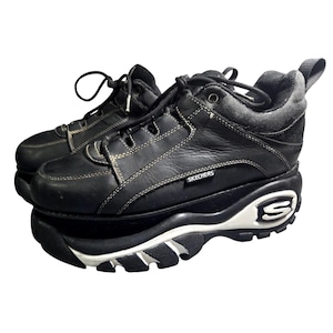 May include: A pair of black leather lace-up sneakers with a thick platform sole. The shoes feature white contrast stitching, a black and white midsole, and a prominent white S logo on the heel. The Skechers brand name is printed on the side.