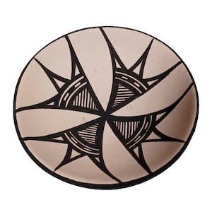 May include: A round, beige ceramic bowl with a black geometric design. The design features a central starburst pattern with radiating lines and triangular shapes. The bowl has a black rim and a smooth surface.