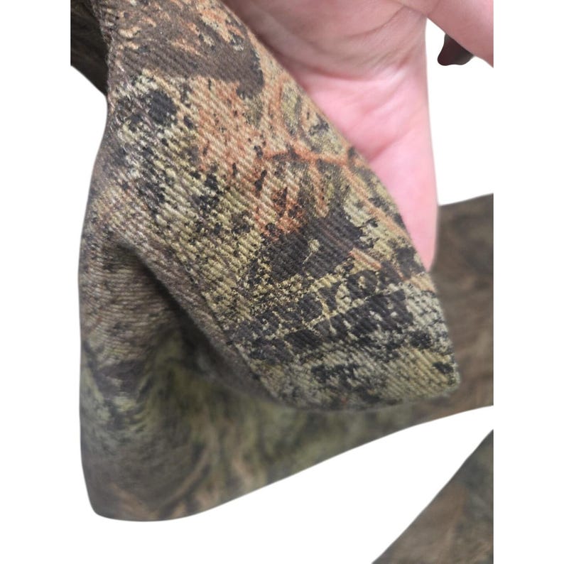 May include: Close-up of a camouflage fabric, featuring a blend of brown, green, and tan hues. The material appears to be a durable textile, possibly for outdoor wear or hunting apparel. The pattern is detailed and intricate.