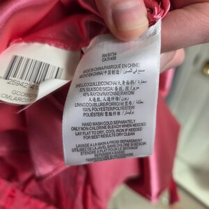May include: Close-up of a clothing tag with text in multiple languages. The tag is white with black text and is attached to a pink fabric. The tag includes care instructions and fabric composition details.