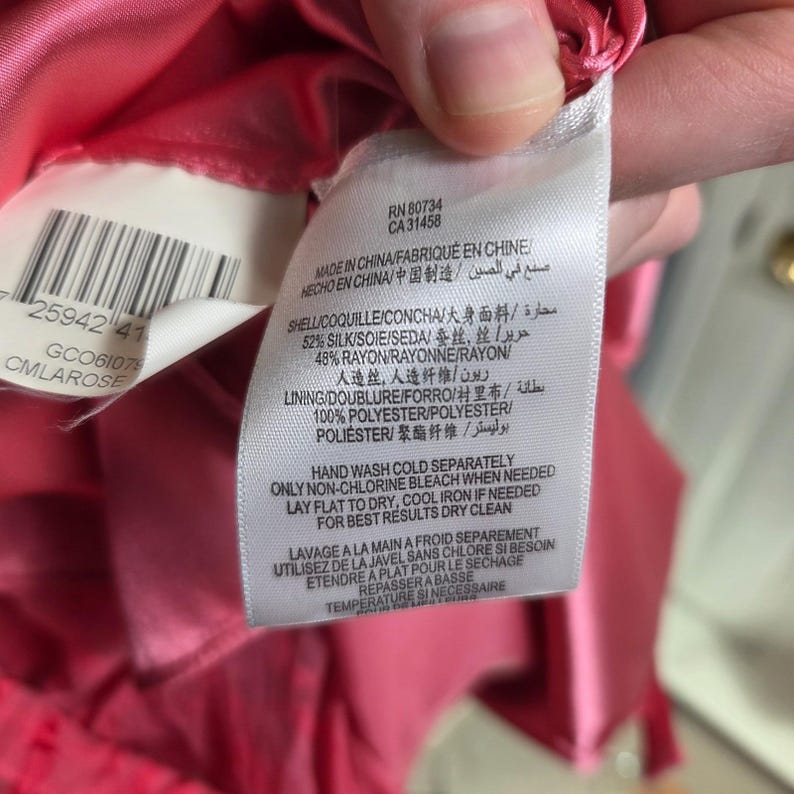 May include: Close-up of a clothing tag with text in multiple languages. The tag is white with black text and is attached to a pink fabric. The tag includes care instructions and fabric composition details.