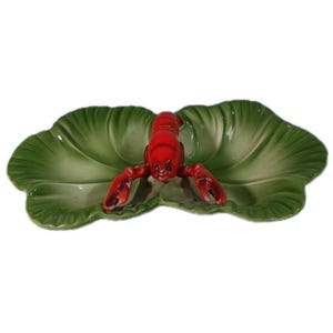 May include: A ceramic serving dish shaped like a green leaf with a bright red lobster figure attached to the center. The dish features a glossy finish and molded details that mimic the veins of a leaf, providing a wide, shallow surface area.