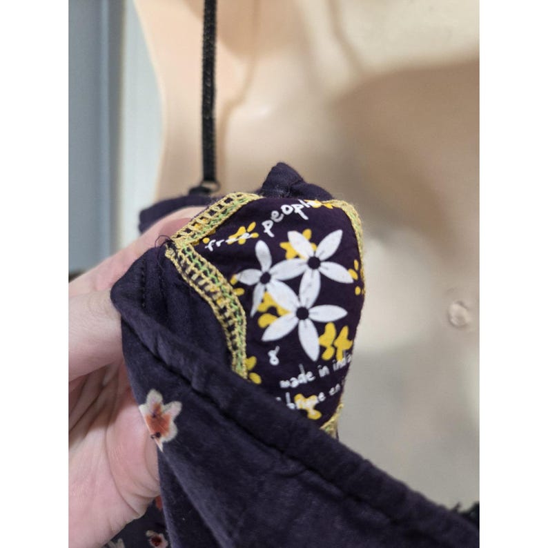Puede incluir: Primer plano de una prenda azul oscuro con estampado floral. La prenda tiene una etiqueta con las palabras "free people" y el texto "made in india". La etiqueta est&aacute; decorada con flores blancas y amarillas y un borde amarillo.