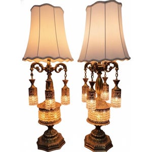 May include: Pair of vintage brass table lamps with white fabric shades. Each lamp features ornate detailing, hanging crystal accents, and a decorative base. The lamps are designed with a classic, elegant aesthetic, suitable for a living room or bedroom.