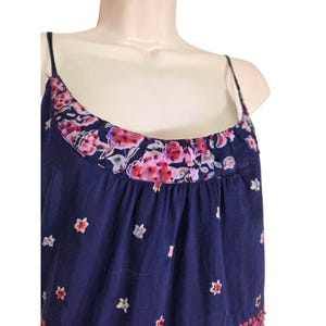 Puede incluir: Un top camisola azul marino con tirantes finos y un escote con estampado floral. El escote presenta una banda de flores rosas y moradas sobre un fondo azul marino. La parte superior est&aacute; decorada con peque&ntilde;os dise&ntilde;os en forma de estrella blanca.