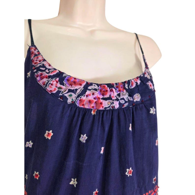 Puede incluir: Un top camisola azul marino con tirantes finos y un escote con estampado floral. El escote presenta una banda de flores rosas y moradas sobre un fondo azul marino. La parte superior est&aacute; decorada con peque&ntilde;os dise&ntilde;os en forma de estrella blanca.