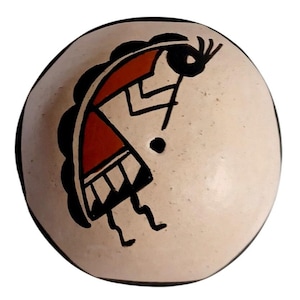 May include: A round, off-white ceramic Kokopelli figure with a black border. The Kokopelli design features a red and black figure with a humpback, playing a flute. The figure is painted on the front of the ceramic piece.