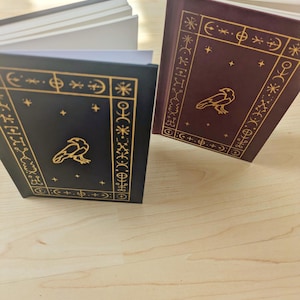 May include: Two hardback books with gold embossed designs and a raven illustration. One book is dark blue, the other is burgundy. Both have gold decorative borders and star accents. The pages are white.