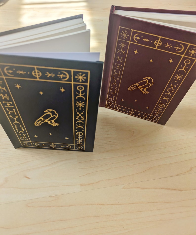 May include: Two hardback books with gold embossed designs and a raven illustration. One book is dark blue, the other is burgundy. Both have gold decorative borders and star accents. The pages are white.
