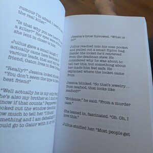 Pu&ograve; includere: Pagine di un libro aperto con testo stampato. Il testo include un dialogo tra i personaggi di nome Julius e Jessica, che discutono di un caso di omicidio e di un medaglione recuperato da uno stufato di morto. Il libro &egrave; aperto su una superficie bianca.