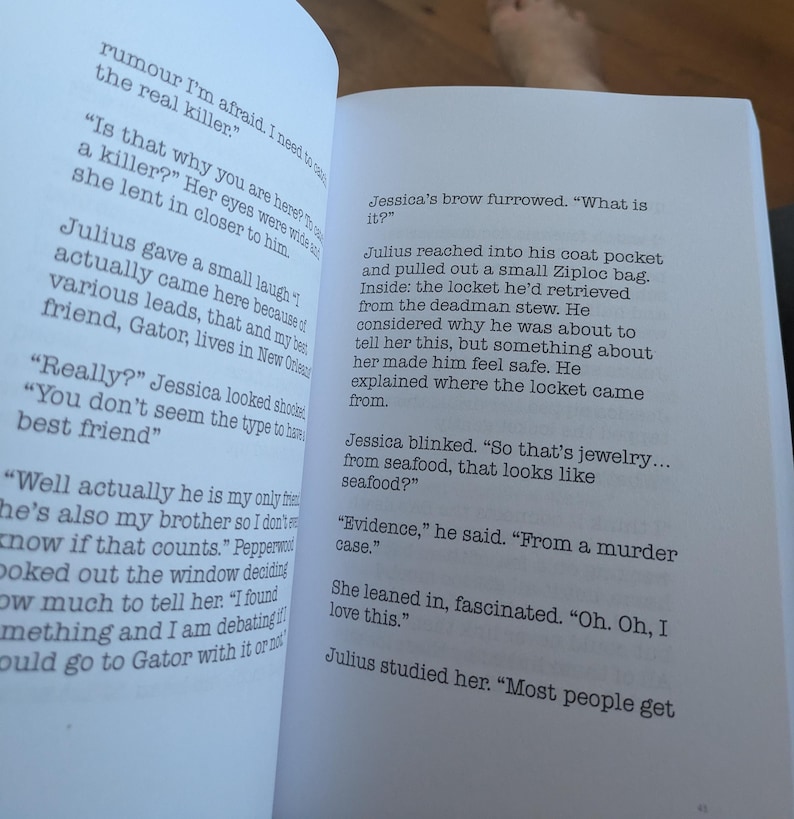 Pu&ograve; includere: Pagine di un libro aperto con testo stampato. Il testo include un dialogo tra i personaggi di nome Julius e Jessica, che discutono di un caso di omicidio e di un medaglione recuperato da uno stufato di morto. Il libro &egrave; aperto su una superficie bianca.
