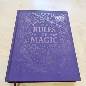 Puede incluir: Un libro de tapa dura de color morado titulado The Rules of Magic con ilustraciones en lámina metálica en la portada. El diseño presenta una bola de cristal, cartas del tarot, una llave, una escoba, una vela y motivos florales.