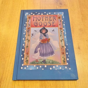 May include: A hardback book titled "MOTHER GOOSE" with a blue cover and white polka dots. The cover illustration shows a woman in a purple dress and a white goose. The book is on a wooden surface.