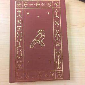 May include: A maroon book with gold foil accents. The cover features a gold raven illustration within a gold border of symbols and stars. The book is on a light wood surface.